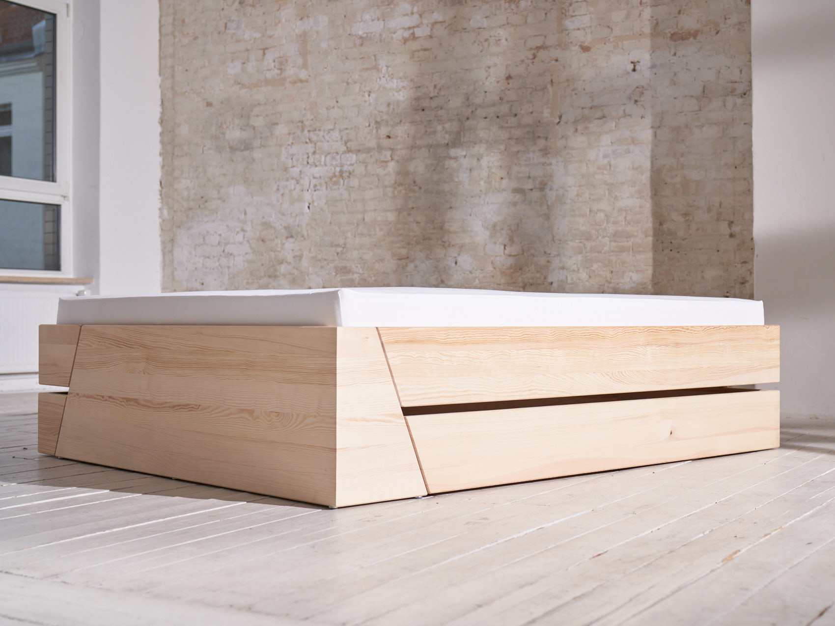 Massivholz Stauraumbett Schublade geschlossen schräge Ansicht auf weißem Holzboden vor weißer Backsteinwand