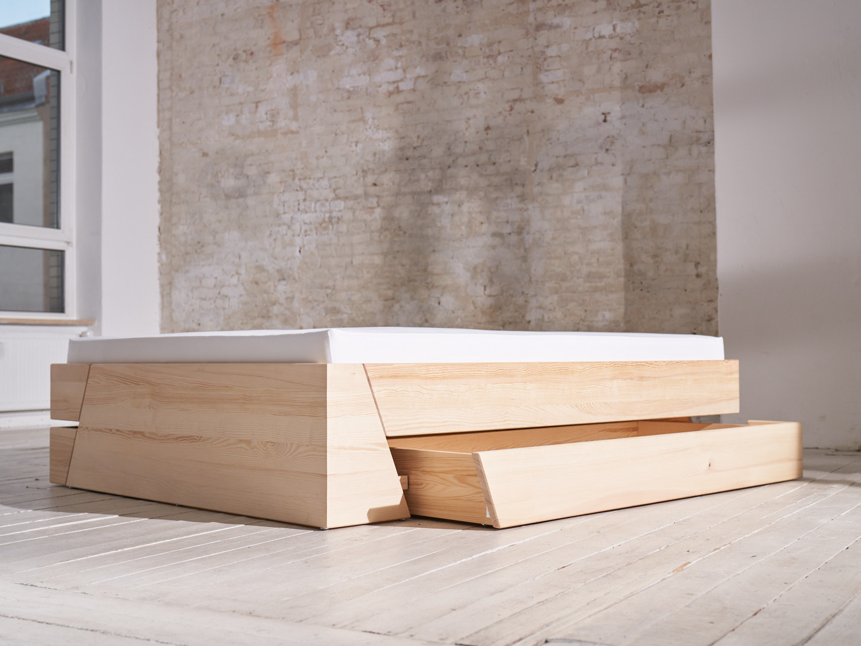 Massivholz Stauraumbett Schublade geöffnet schräge Ansicht auf weißem Holzboden vor weißer Backsteinwand